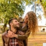 A smiling woman with curly hair playfully hugs a bearded man from behind in a park. Both wear brown jackets and look at each other happily, surrounded by trees and sunlight.