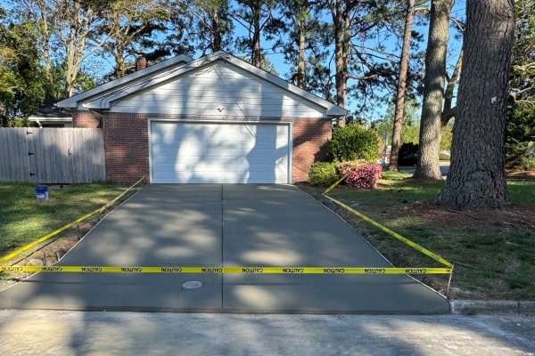 A newly poured concrete driveway in front of a house, blocked off with caution tape for drying. Surrounding trees and shrubs visible.