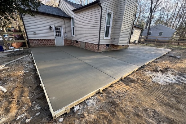 Newly poured concrete patio beside a house, with wooden framing and surrounding dirt area. Trees are visible in the background.