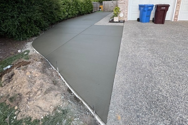 Freshly poured concrete driveway with a gravel edge, next to a textured concrete surface and trash bins in the background.