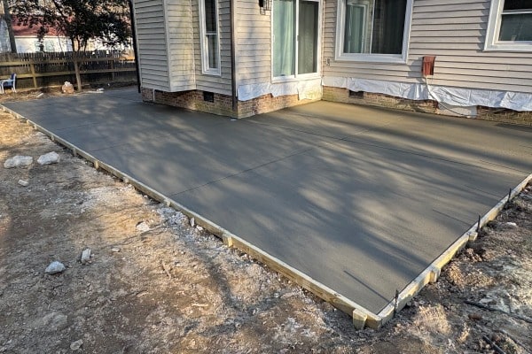 Freshly poured concrete slab next to a house, with wooden forms along the edges and shadowed areas in the yard.