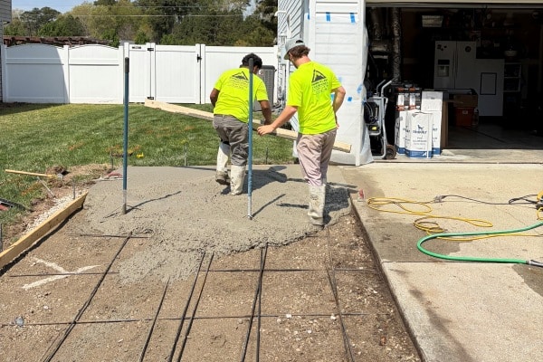 Two workers in bright yellow shirts are pouring concrete onto a gravel surface with rebar framing.