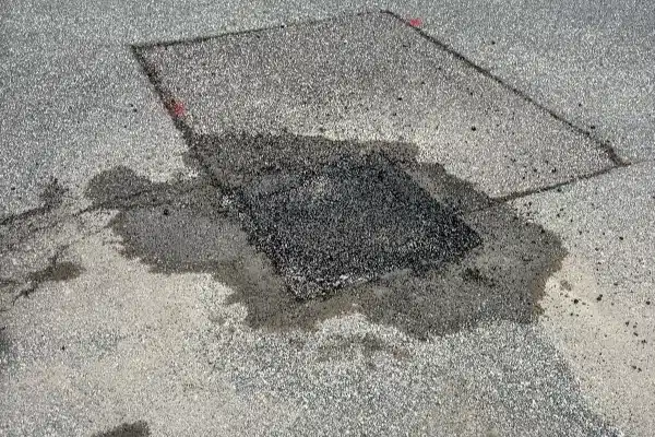 A dark manhole cover surrounded by a puddle of water on an asphalt surface, with some gravel scattered nearby.