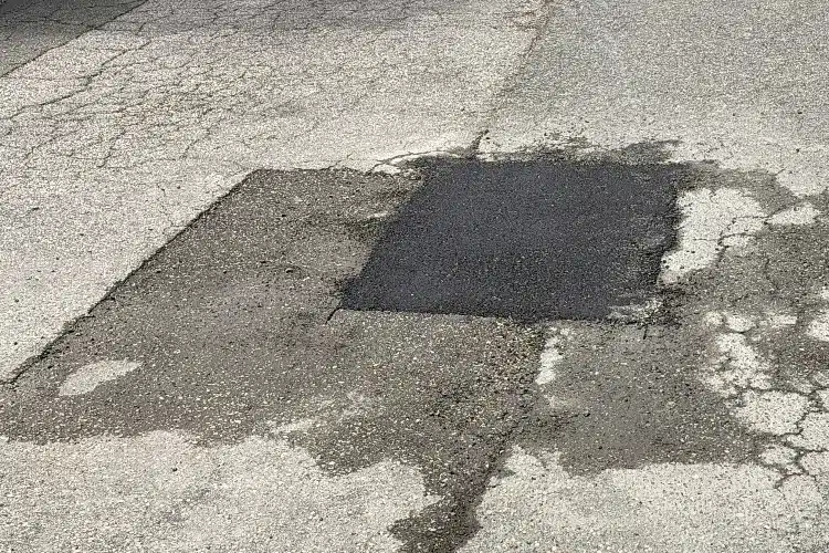 Pavement with a rectangular asphalt patch, surrounded by cracked concrete and gravel.