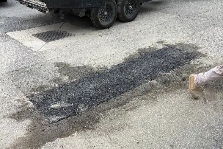A patch of fresh asphalt on a roadway, with wet areas around it and a person walking past in light-colored pants.