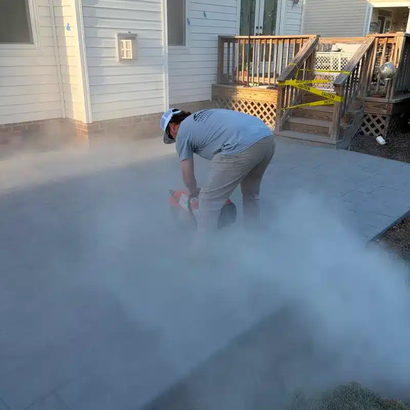 A person uses a cutting tool on a stone patio, creating a cloud of dust in a residential area. Caution tape is visible nearby.