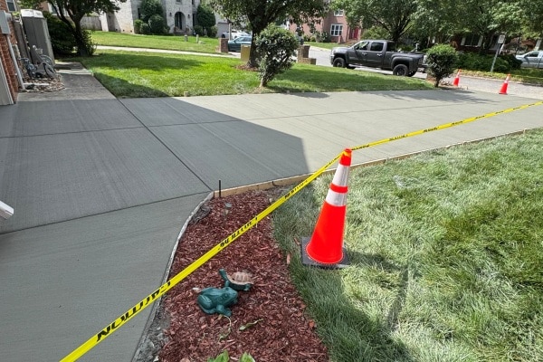 Freshly poured concrete driveway with caution tape and traffic cones; decorative landscape features in foreground.