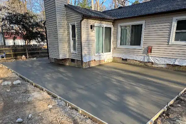 Concrete patio in front of a house under construction, with light-colored siding, windows, and a clear sky in the background.