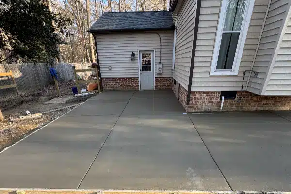 A newly poured concrete patio is adjacent to a house with light-colored siding and a door, surrounded by trees.