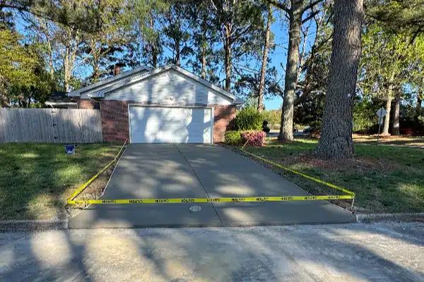 Newly poured concrete driveway in front of a house, marked with caution tape, surrounded by trees and grass.