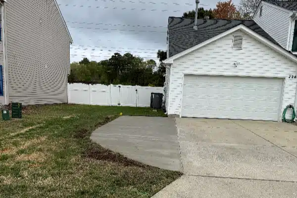 A concrete driveway leads to a white garage; green grass and a fenced yard are visible in the background under a cloudy sky.