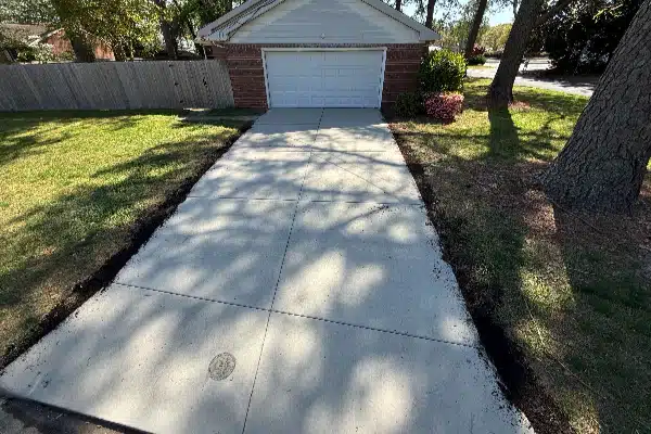Driveway leading to a garage, bordered by grass and mulch, with trees on the right side and a pink bush in the background.