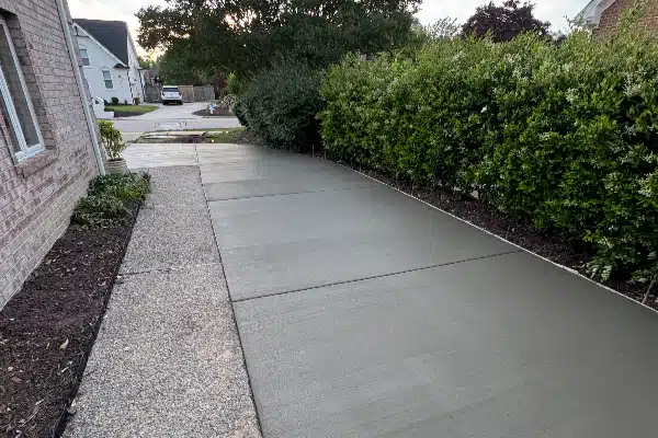 Concrete path leading from a brick house, bordered by shrubs and mulch, with a glimpse of a driveway in the background.