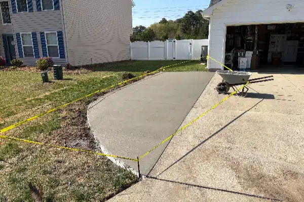Freshly poured concrete pathway, marked off with yellow caution tape, next to a garage and a wheelbarrow.