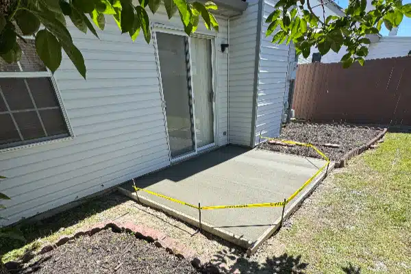 Concrete patio area with yellow caution tape around the perimeter, adjacent to a house and landscaped garden beds.