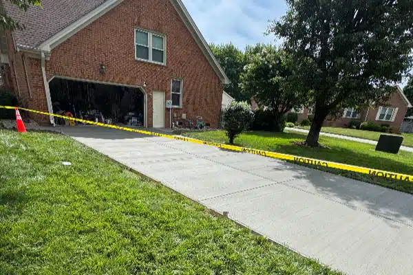 A residential driveway with caution tape across it, adjacent to a brick house and grassy yard.