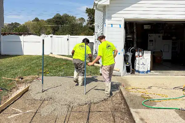 Two workers in bright yellow shirts are pouring concrete near a house, with rebar frames standing in the fresh concrete.