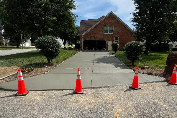 Freshly poured concrete driveway with orange traffic cones marking the area, surrounded by green grass and trees.