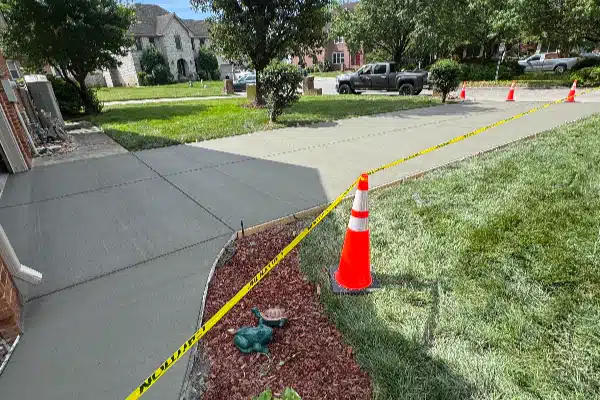 Newly poured concrete driveway with traffic cones and caution tape surrounding the area, grass and shrubs in the foreground.