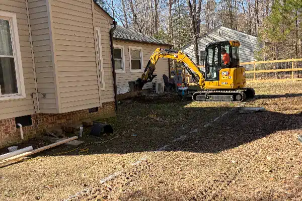 Excavator positioned near the side of a house, working in a yard with trees and equipment visible in the background.