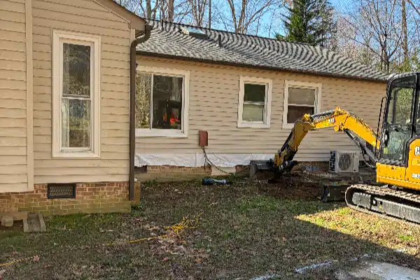 Construction equipment digging near the side of a house, with a clear blue sky and trees in the background.