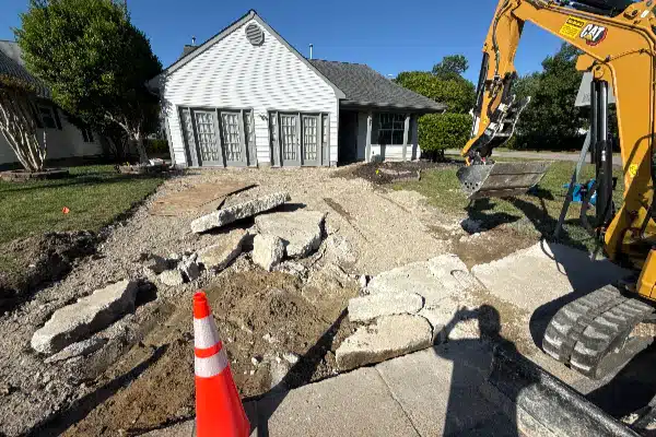 Construction site with broken concrete slabs, a pile of dirt, a construction vehicle, and a house in the background.