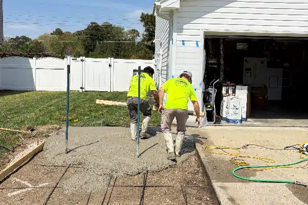 Two workers in bright yellow shirts are pouring concrete in a backyard. Tools and equipment are nearby.