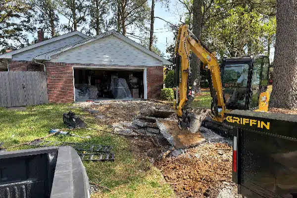 A construction site with an excavator digging by a garage. Debris and gravel are scattered around the work area.