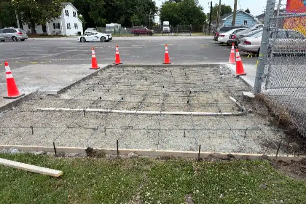 A construction site showing a gravel base with rebar grid, surrounded by orange traffic cones and a fenced area.