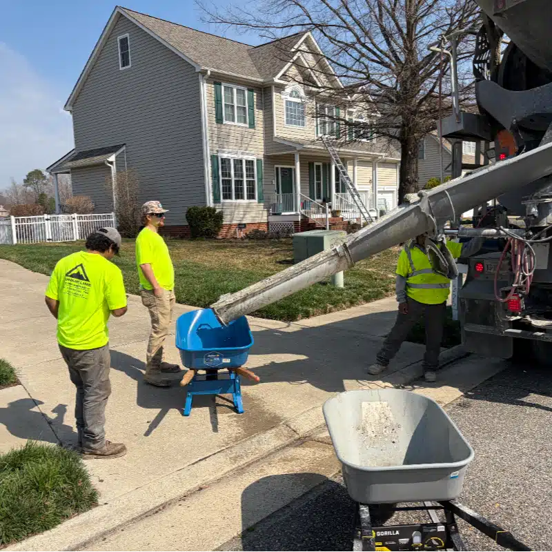 Construction workers in bright shirts operate a concrete mixer pouring into a wheelbarrow on a residential sidewalk.