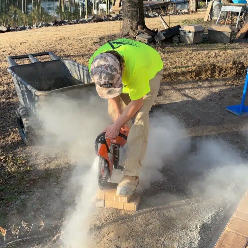 A person in a bright yellow shirt uses a saw on a brick, creating dust in an outdoor setting. A wheelbarrow is nearby.