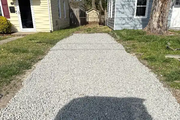 Gravel driveway between two houses, with grass on either side and a tree in the background.