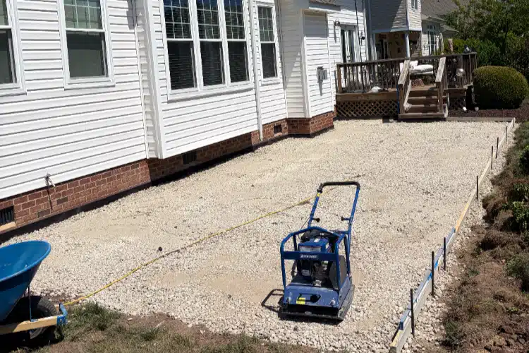A gravel surface is prepared for paving next to a house, with a compactor and a wheelbarrow nearby.