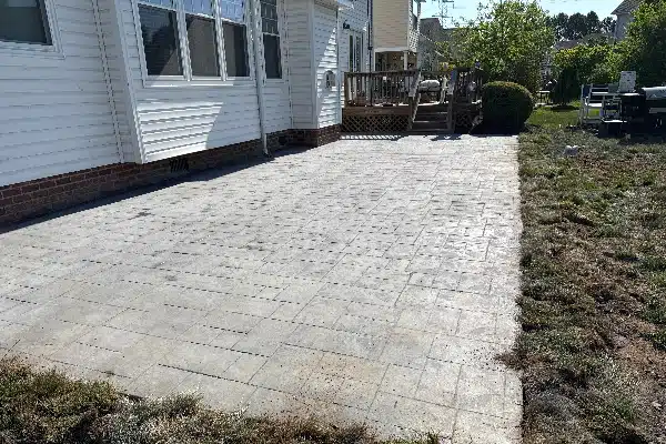 A paved patio area beside a house, with a wooden deck and green grass surrounding it.