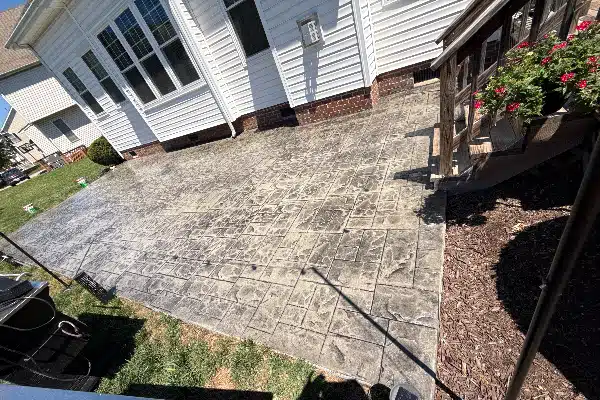 A stamped concrete patio is shown beside a white house, with a wooden railing and flowers in a planter on the right.