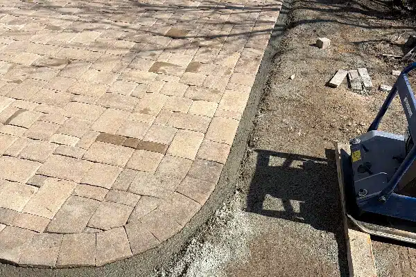 Paved area with light-colored stones, partially finished, next to grey wet cement, and compacting machine nearby.