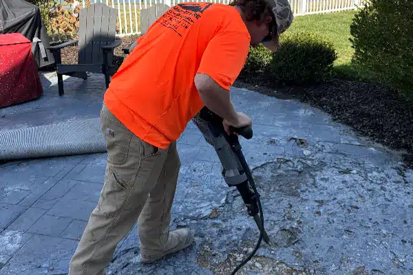 A person in an orange shirt uses a tool to break up a concrete surface outside, surrounded by grass and patio furniture.