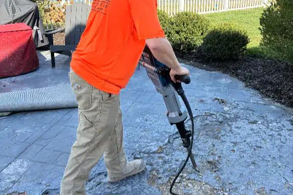 A person in an orange shirt uses a jackhammer on a concrete patio, with a yard and furniture in the background.