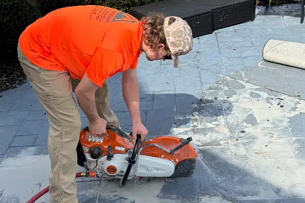 A person in an orange shirt operates a concrete saw on a stone surface, with dust and debris visible.