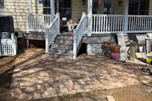 Deck stairs lead to a paved patio area with scattered tools and planters, bordered by a house and grass.