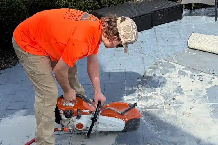 A person in an orange shirt operates a concrete saw on a stone patio, with dust and debris around.